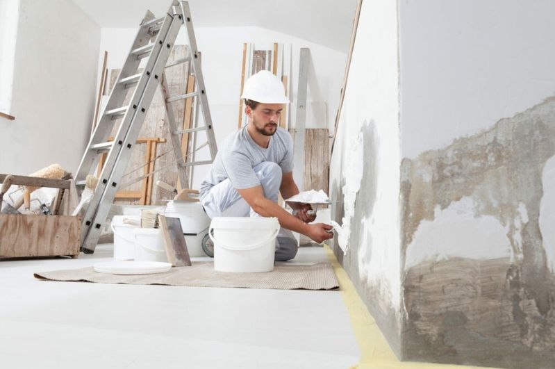 Handwerker mit Schutzhelm kniet auf Boden und trägt Spachtelmasse an Wand auf, im Hintergrund Leiter und Baumaterialien