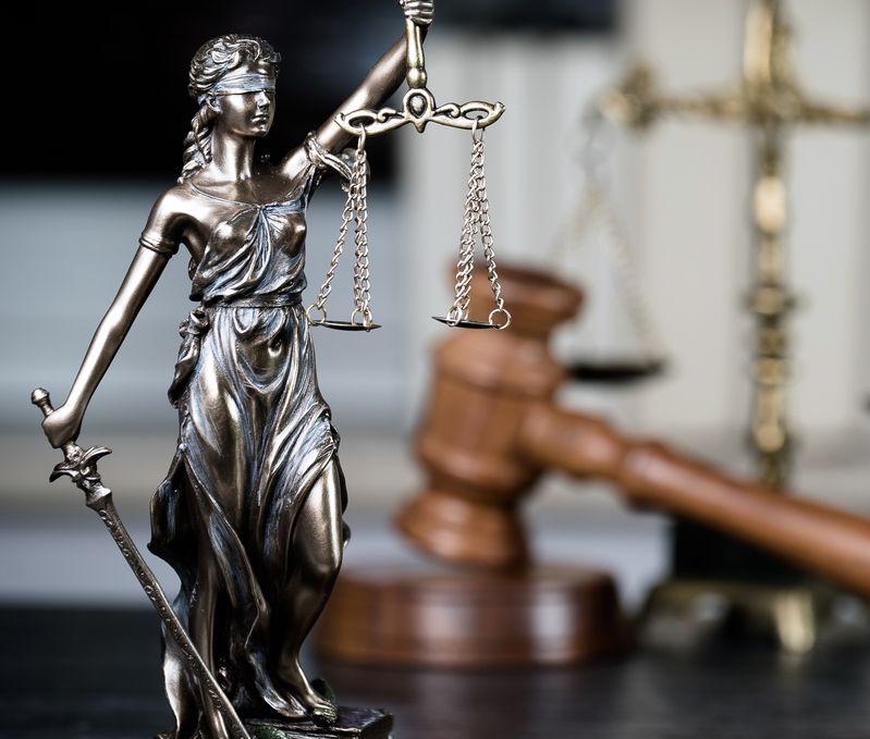 Bronzefigur der Justitia mit Waage und Schwert vor einem Holzrichterhammer und einer unscharfen Justitia-Statue im Hintergrund