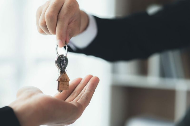 Eine Hand übergibt einer anderen Hand einen Schlüsselbund mit einem Schlüsselanhänger in Form eines Hauses mit der Aufschrift 'Home'